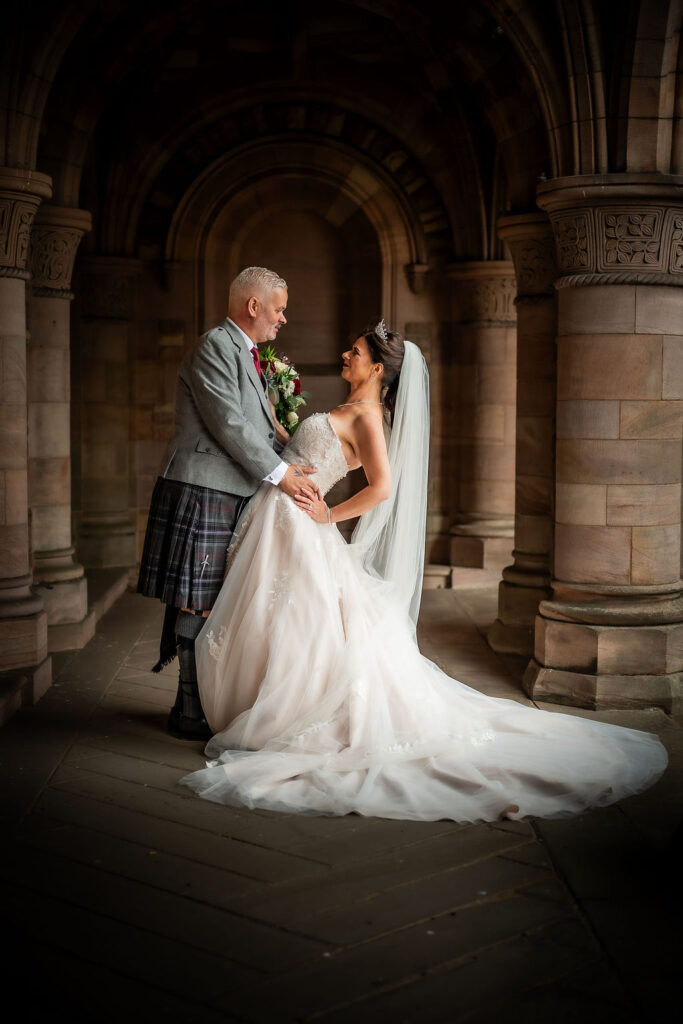 Bride and groom gazing at each other beneath a grand stone archway, with the bride's flowing white gown and veil contrasting against the warm historic architecture in a romantic wedding portrait.