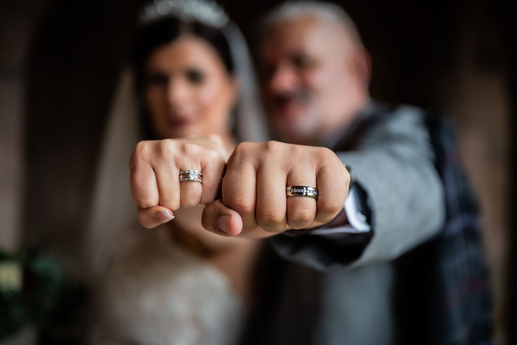 Close-up of the bride and groom holding out their hands to showcase their wedding rings, with the couple softly blurred in the background for a creative wedding portrait.