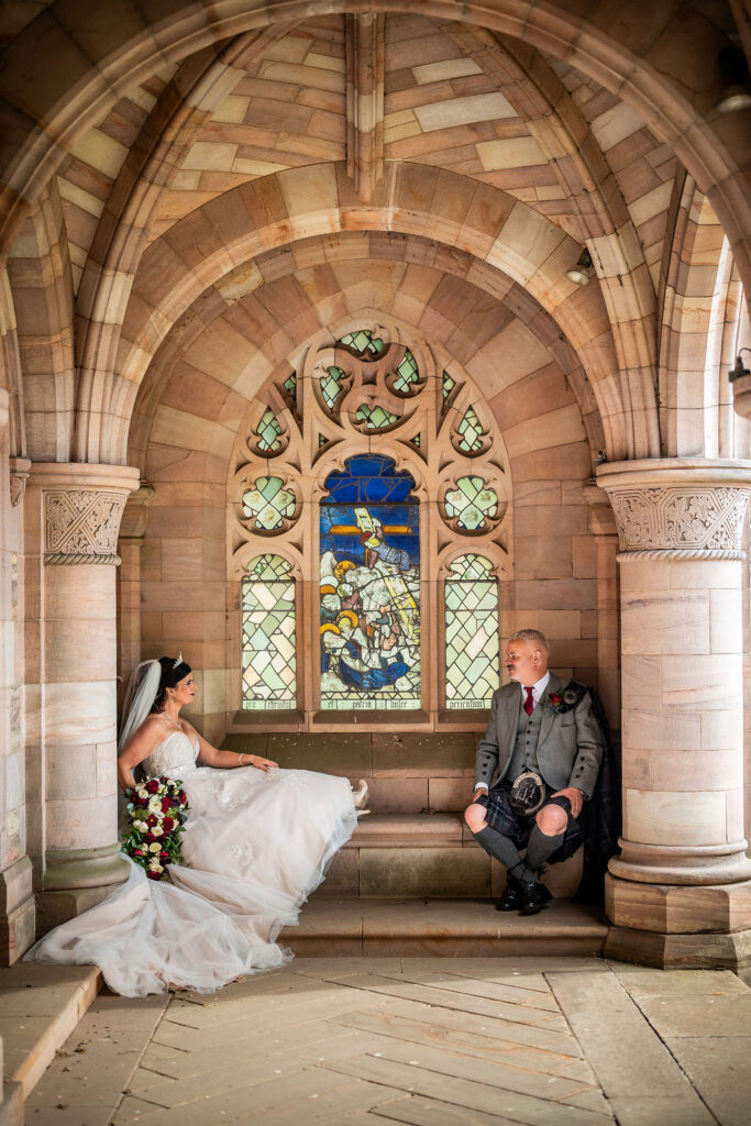 Bride and groom seated beneath an ornate sandstone archway inside a historic building, facing each other with a stained-glass window as the backdrop on their wedding day.