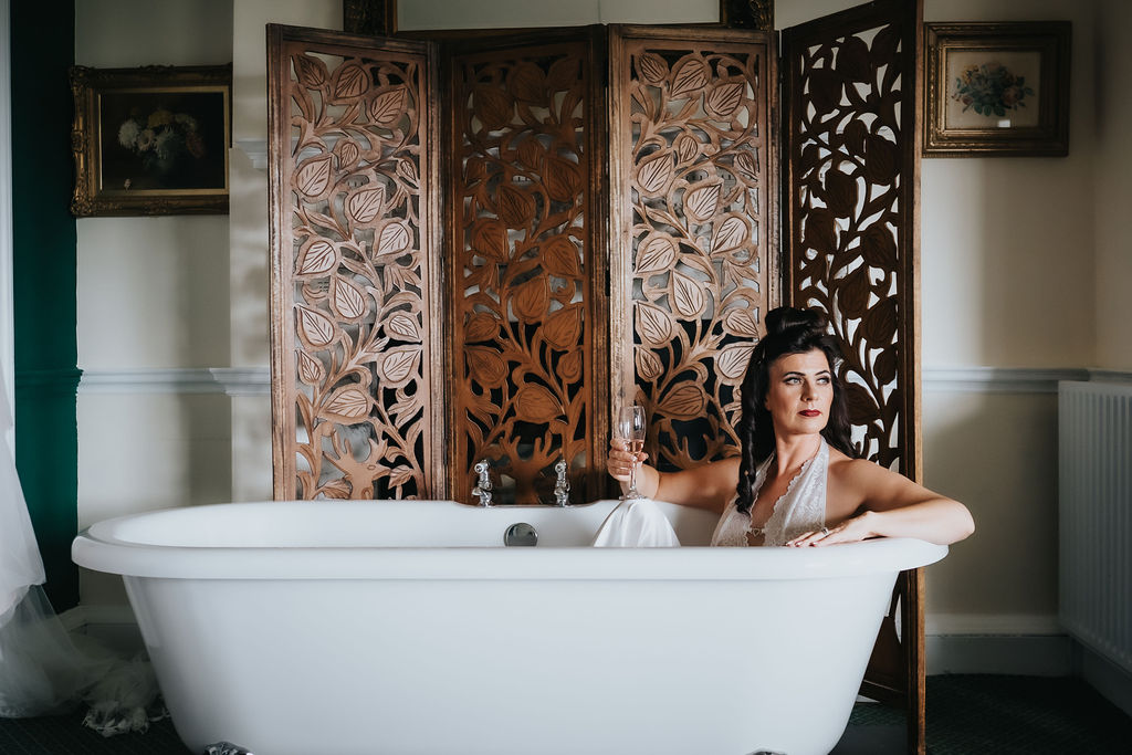 Bride in a white wedding gown sitting gracefully in a freestanding bathtub, posing in front of an ornate carved wooden room divider during a stylish bridal portrait indoors.