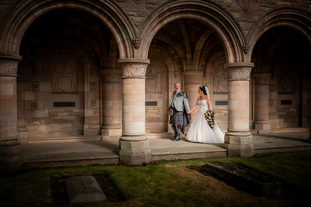 Bride in a white wedding gown and groom wearing a traditional Scottish kilt hold hands beneath a row of historic stone arches, creating a timeless wedding portrait in a grand courtyard.