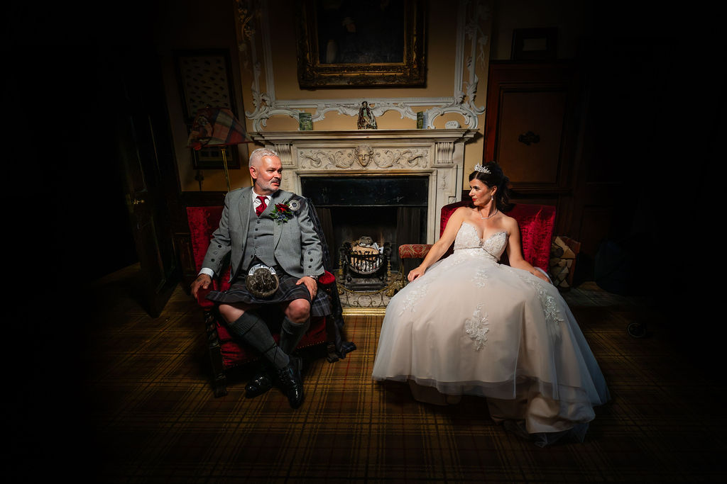 Bride in a white wedding gown and groom in a traditional Scottish kilt seated in separate red armchairs across from each other in front of an elegant fireplace, sharing a smile in a warmly lit historic room.