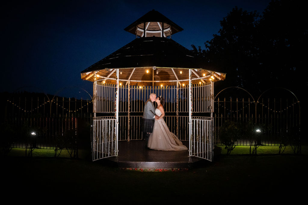 Bride and groom embracing inside a beautifully lit white gazebo at night, surrounded by glowing string lights in a romantic outdoor wedding portrait.