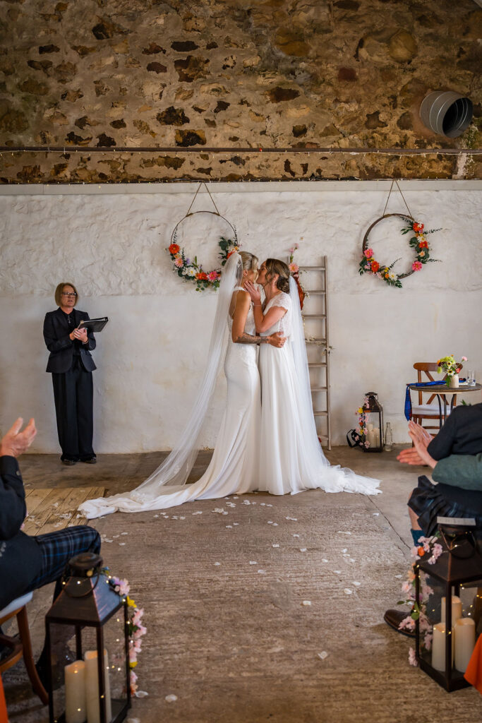 Two brides share their first kiss after exchanging vows during a rustic indoor wedding ceremony, surrounded by floral decorations as guests applaud.