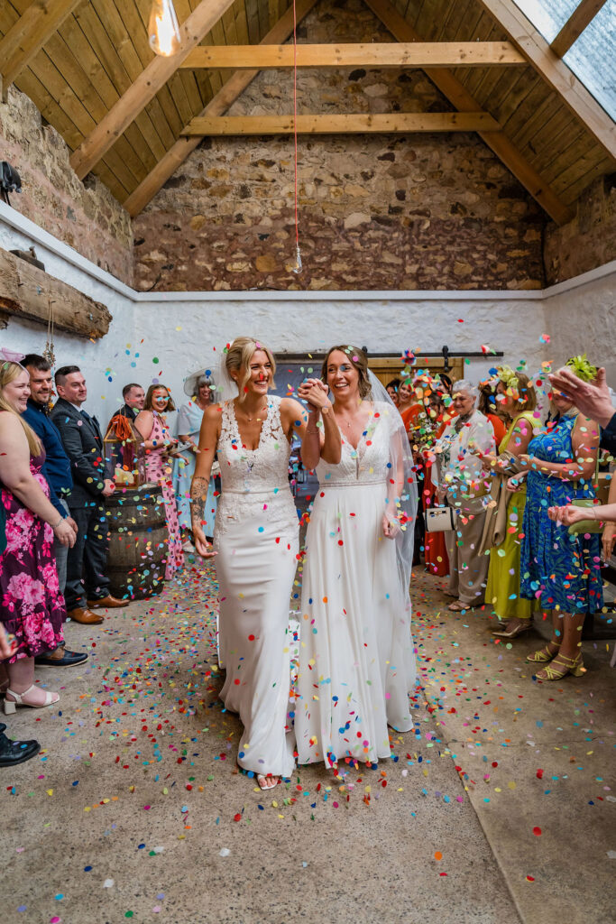 Two brides walk hand in hand through a colorful confetti shower inside a rustic wedding venue, smiling as family and friends celebrate around them.
