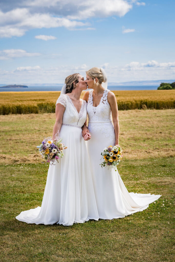 Two brides in elegant white wedding dresses share a kiss while holding hands in a sunlit countryside field, with colorful bouquets, blue skies, and a scenic coastal backdrop.