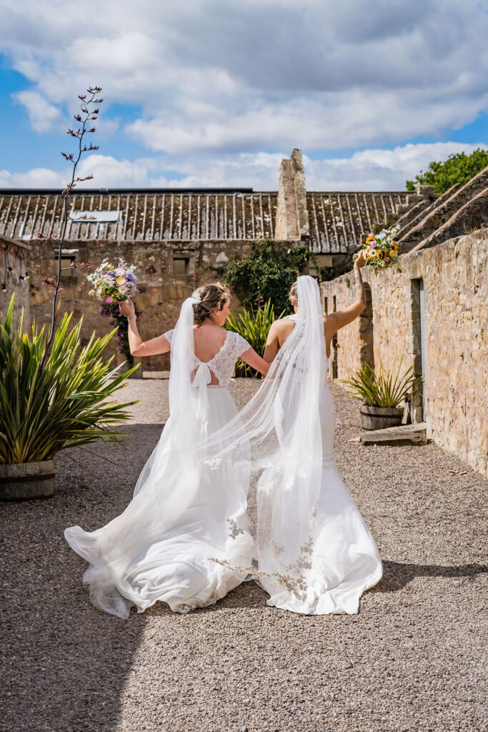 Two brides in flowing white wedding gowns walk hand in hand through a rustic stone courtyard, raising colorful bouquets as their veils trail behind on a bright wedding day.