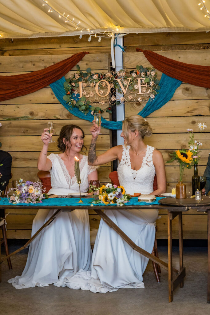 Two brides raise champagne glasses in a joyful wedding toast at their reception, seated beneath a floral "LOVE" sign with rustic décor and colorful bouquets.