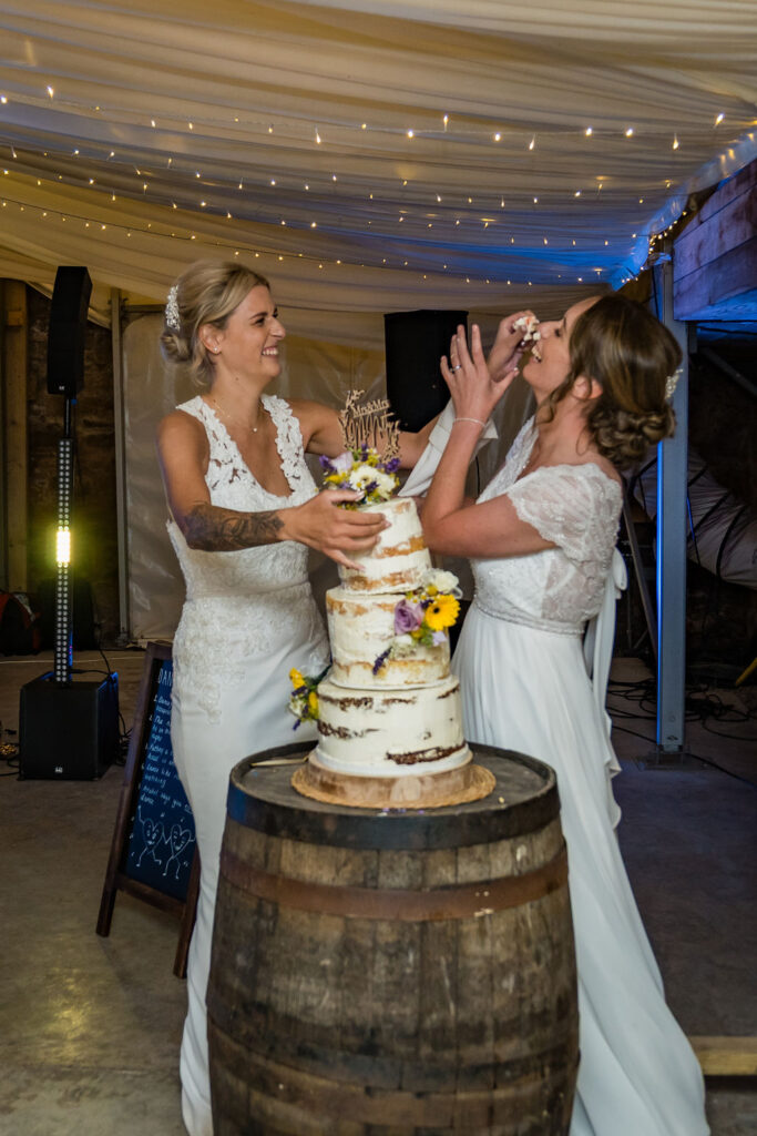 Two brides laugh as they playfully feed each other wedding cake beside a rustic three-tier cake on a wooden barrel during their reception under twinkling fairy lights.