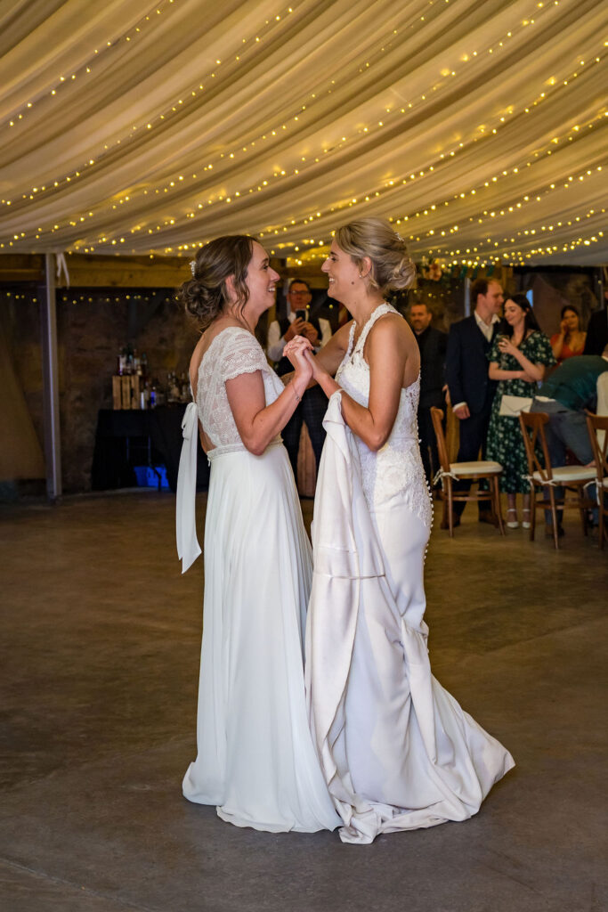Two brides share their first dance at their wedding reception, smiling and holding hands beneath warm string lights while guests watch in the background.