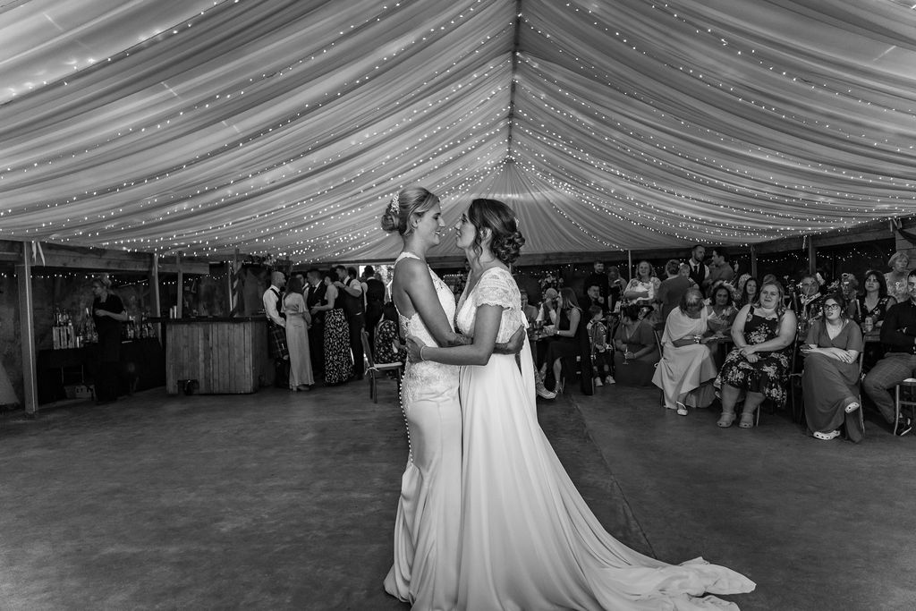 Black-and-white photo of two brides sharing their first dance beneath twinkling fairy lights at their wedding reception, surrounded by family and friends.