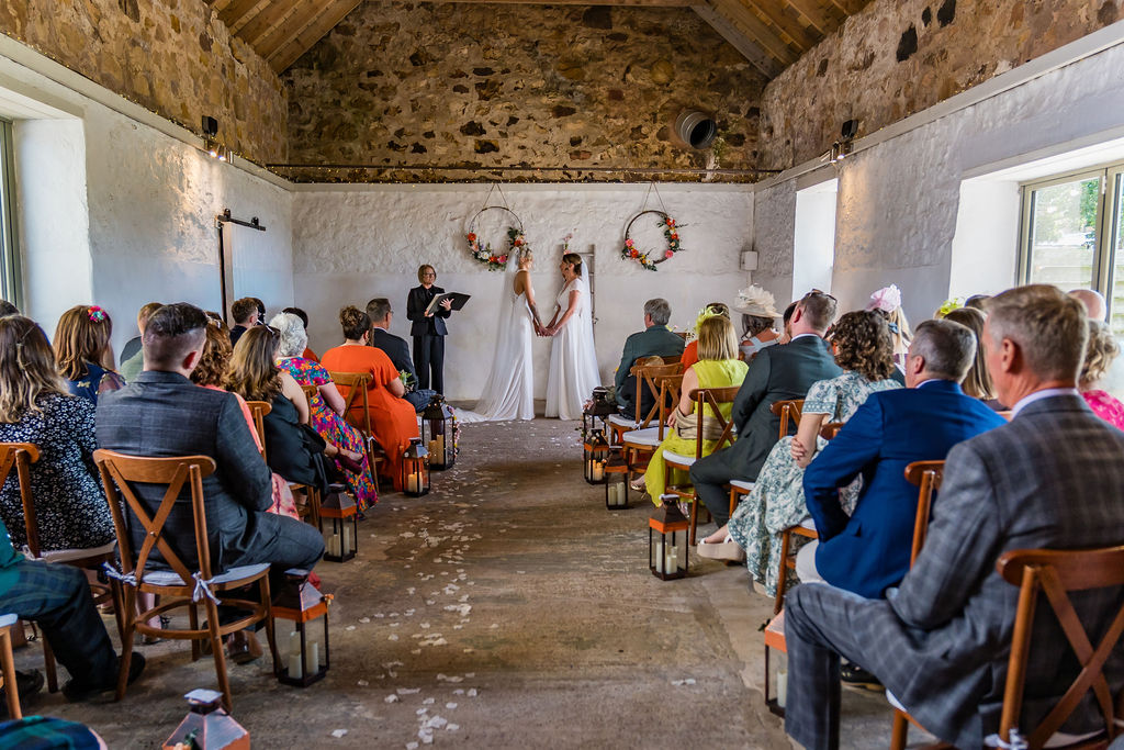 Guests watch as two brides stand hand in hand during their wedding ceremony inside a rustic stone barn, with floral wreaths, lanterns, and a petal-lined aisle.