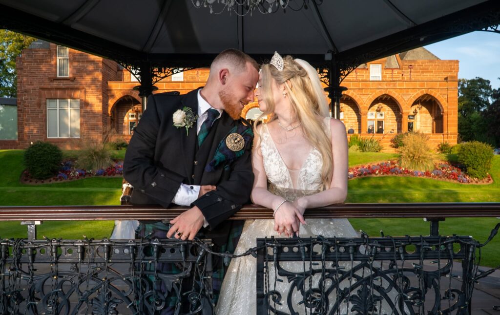 Bride and groom sharing a romantic forehead-to-forehead moment under a decorative gazebo on their wedding day. The groom wears a traditional Scottish kilt with a tartan tie and boutonnière, while the bride wears a sparkling lace wedding gown, tiara, and veil. A red sandstone venue and landscaped gardens glow in warm golden-hour light in the background.