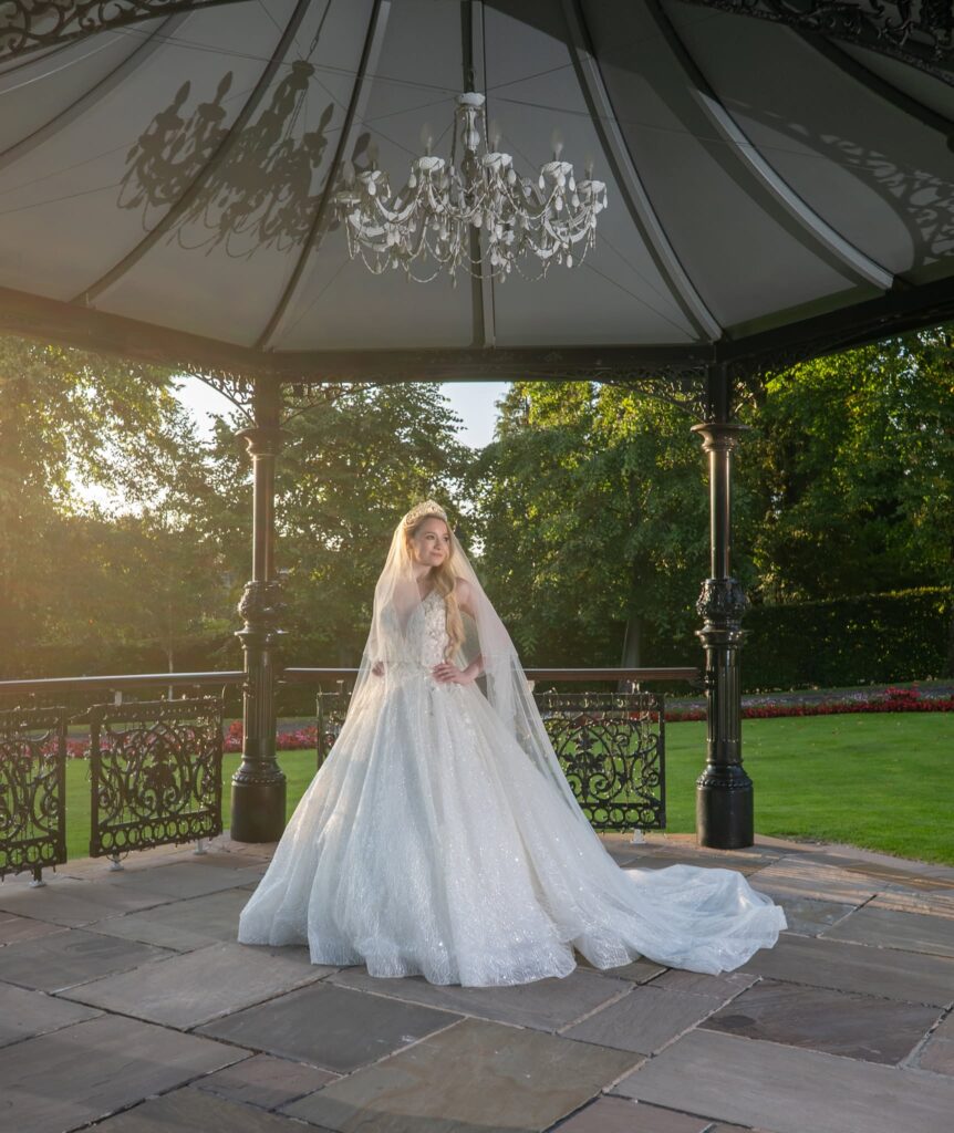 Bride standing beneath an elegant wrought-iron gazebo during golden hour, wearing a sparkling ballgown wedding dress with a long train, veil, and tiara. Warm sunlight filters through the surrounding gardens, while a crystal chandelier overhead adds a romantic touch to this timeless outdoor bridal portrait.