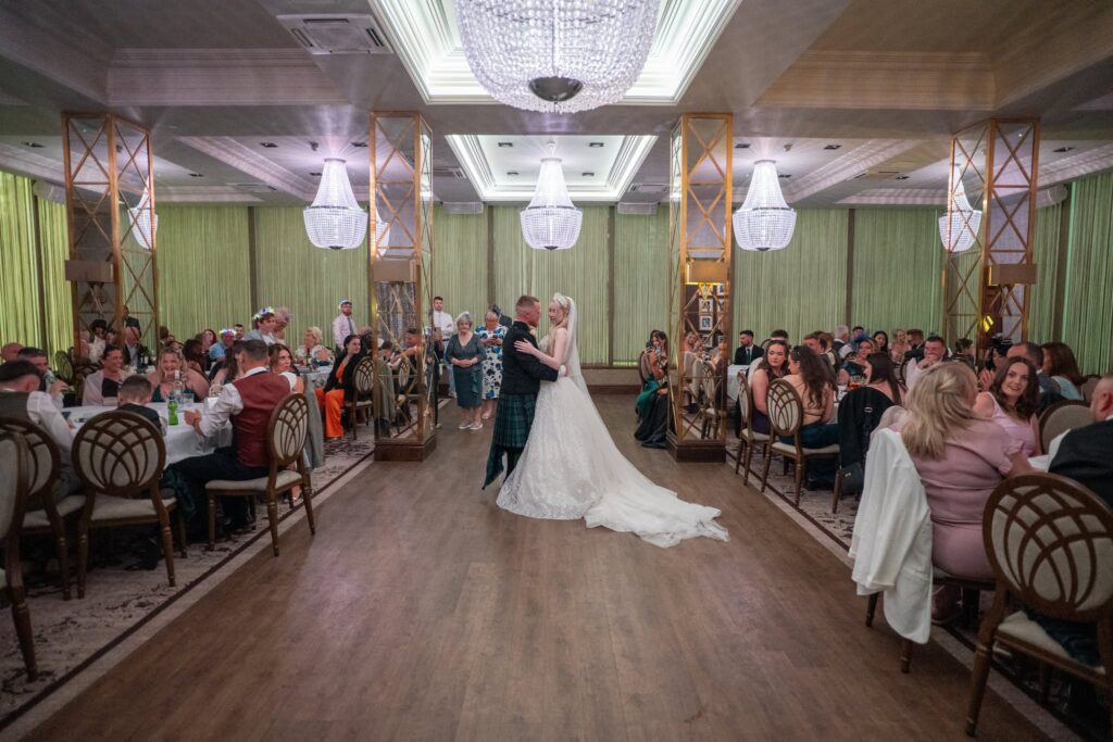 Bride and groom sharing their first dance in the centre of an elegant wedding reception. The bride wears a sparkling lace wedding gown with a long train and veil, while the groom wears a traditional Scottish kilt. Family and friends are seated around the dance floor watching the couple beneath grand crystal chandeliers in a beautifully decorated ballroom.