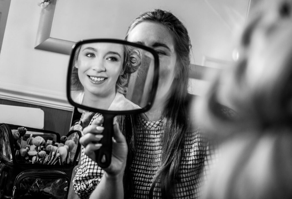 Black and white wedding preparation photo of a bride smiling as she sees her hair and makeup reflected in a handheld mirror. A bridesmaid holds the mirror while makeup brushes and beauty products sit nearby, capturing a candid getting-ready moment before the wedding ceremony.