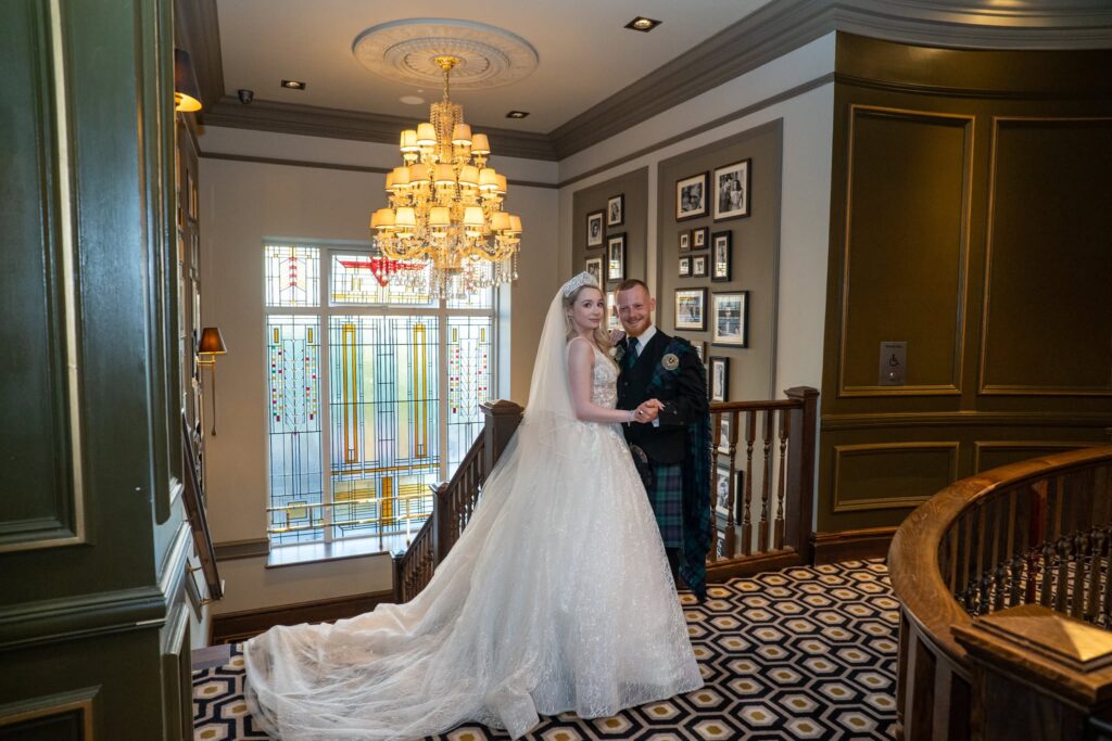 Bride and groom posing together on an elegant staircase inside their wedding venue. The bride wears a sparkling lace wedding gown with a long flowing train, veil, and tiara, while the groom wears a traditional Scottish kilt with tartan, jacket, and sporran. They smile as they hold hands beneath a crystal chandelier, with stained-glass windows and classic décor creating a timeless wedding setting.