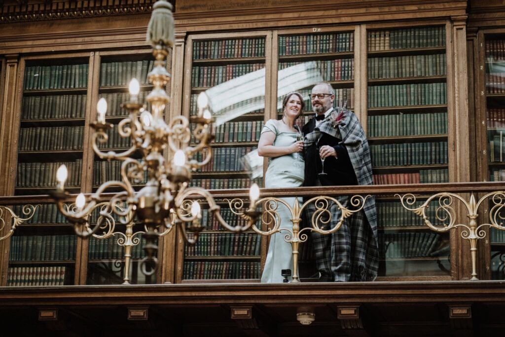 Newlyweds Alan and Gillian share a quiet moment with champagne on the balcony of the Royal College of Physicians of Edinburgh, framed by historic library shelves and an elegant chandelier.