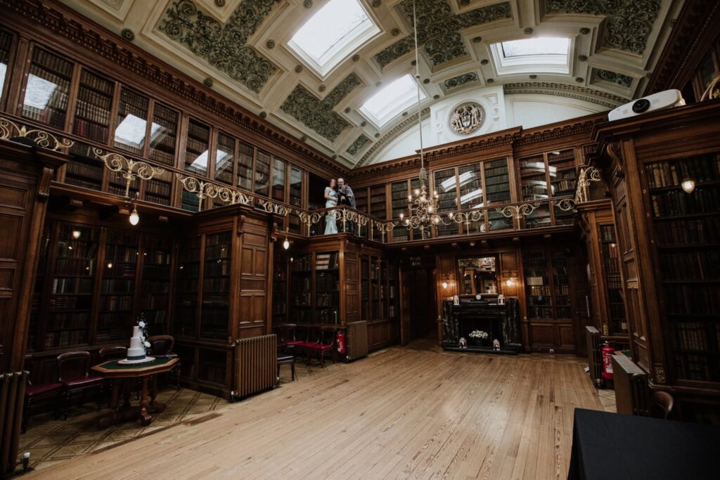 Alan and Gillian pose together on the historic library balcony at the Royal College of Physicians of Edinburgh, surrounded by soaring bookshelves, ornate ceilings, and elegant architecture during their wedding day.