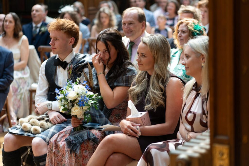 Wedding guests become emotional during Alan and Gillian’s ceremony at the Royal College of Physicians of Edinburgh, as a bridesmaid wipes away tears while holding a bouquet and family members watch the heartfelt vows.