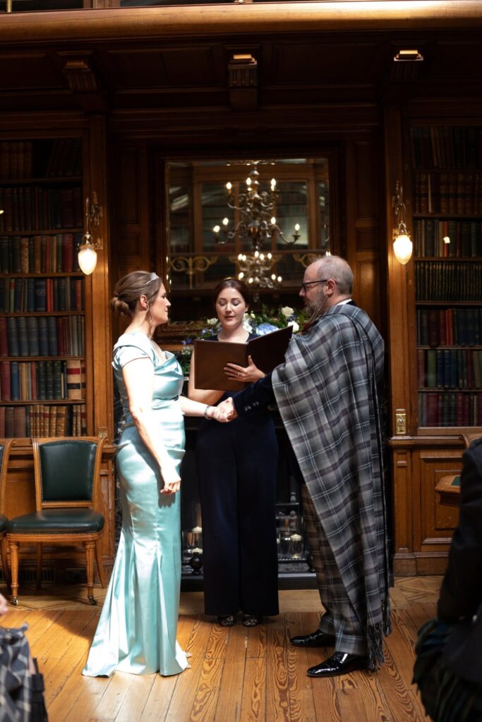Bride and groom hold hands as they exchange vows during their intimate wedding ceremony in the historic New Library at the Royal College of Physicians of Edinburgh, with their celebrant officiating in front of a grand fireplace and floor-to-ceiling bookshelves.
