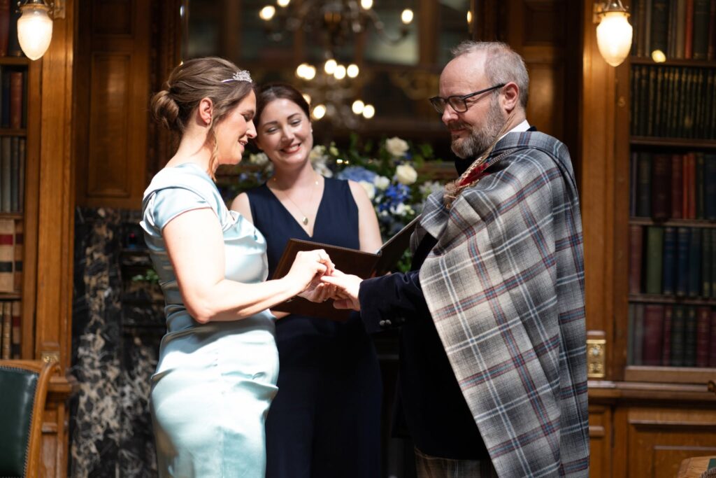 Alan places a wedding ring on Gillian’s finger during their intimate ceremony at the Royal College of Physicians of Edinburgh, as their celebrant smiles in the background surrounded by elegant library interiors.