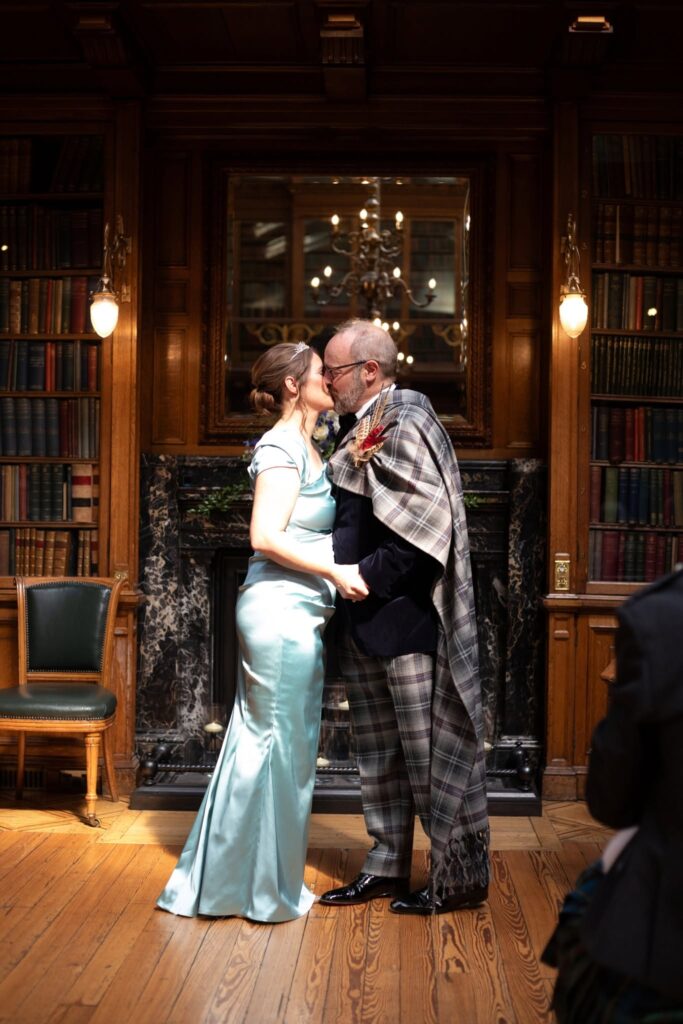 Bride and groom share a kiss in the elegant library of the Royal College of Physicians of Edinburgh, standing in front of a grand fireplace surrounded by historic bookshelves during their intimate wedding ceremony.