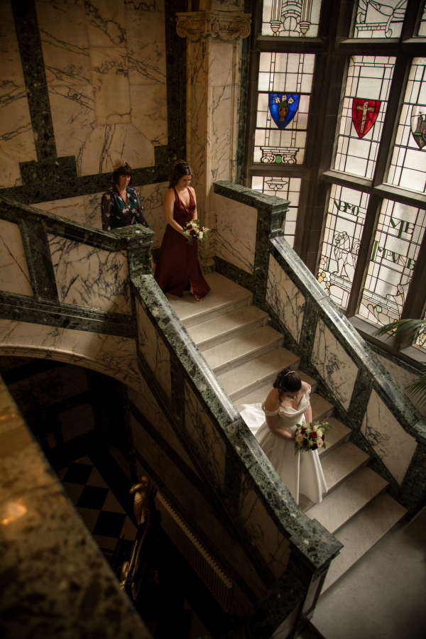 The bride makes her way down an elegant marble staircase, holding a bouquet of white and burgundy flowers, while her bridesmaid and a family member follow behind. Framed by ornate stained-glass windows and grand stone architecture, the scene captures a quiet moment before the wedding ceremony.