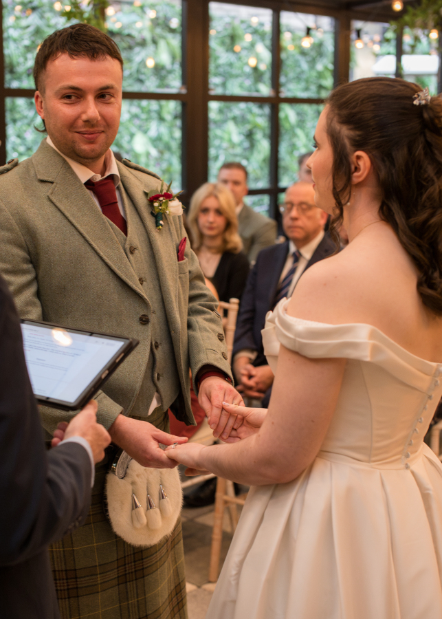 The bride and groom hold hands and exchange rings during their wedding ceremony, sharing a heartfelt moment as they look at one another. The bride wears an elegant off-the-shoulder white gown, while the groom wears a traditional Scottish tartan kilt with a tweed jacket. Family and friends watch from their seats as the celebrant leads the ceremony.