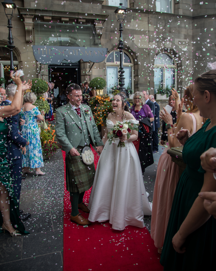 The newlyweds walk hand in hand along a red carpet outside The Scotsman Hotel as guests shower them with colourful confetti. The bride wears an elegant off-the-shoulder white gown and carries a bouquet of white and burgundy flowers, while the groom wears a traditional Scottish tartan kilt with a sage green tweed jacket. Their joyful expressions capture the excitement of celebrating with family and friends after the ceremony.