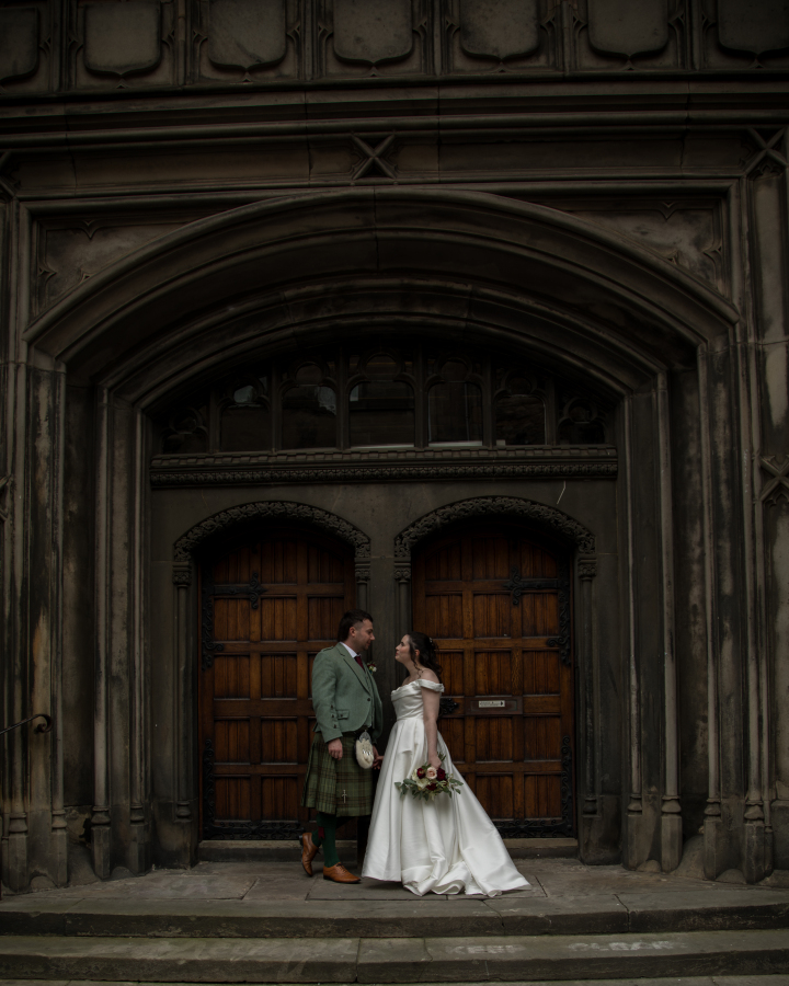 The bride and groom stand together outside a grand historic building, smiling at one another beneath a large stone archway. The bride wears an elegant off-the-shoulder white gown and holds a bouquet of white and burgundy flowers, while the groom wears a traditional Scottish tartan kilt with a sage green tweed jacket. The imposing wooden doors and Gothic stonework create a timeless backdrop for their wedding portrait.