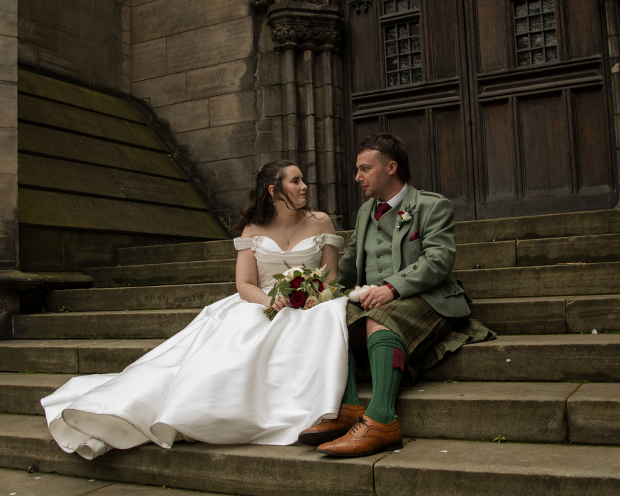 The bride and groom sit together on the stone steps outside a historic building, sharing a quiet moment as they look into each other's eyes. The bride wears an elegant off-the-shoulder white gown and holds a bouquet of white and burgundy flowers, while the groom wears a sage green tweed jacket with a traditional Scottish tartan kilt. The grand stone architecture provides a timeless backdrop for their romantic wedding portrait.