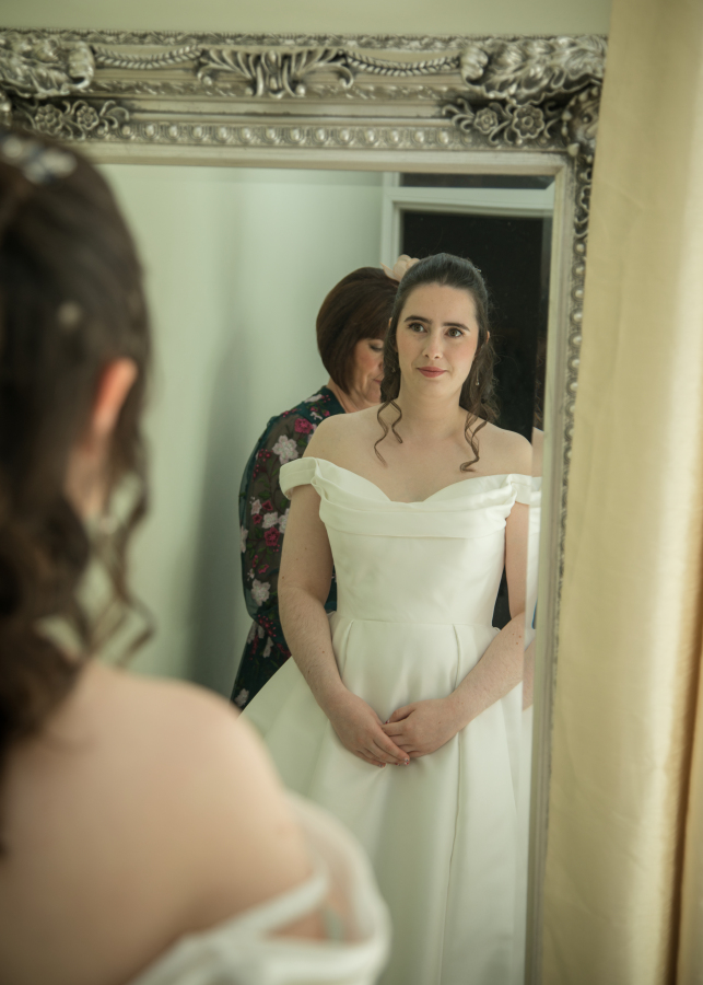 The bride admires her reflection in a mirror while getting ready for her wedding ceremony. She wears an elegant off-the-shoulder white gown as a family member helps with the final preparations behind her, capturing a quiet and emotional moment before the celebrations begin.