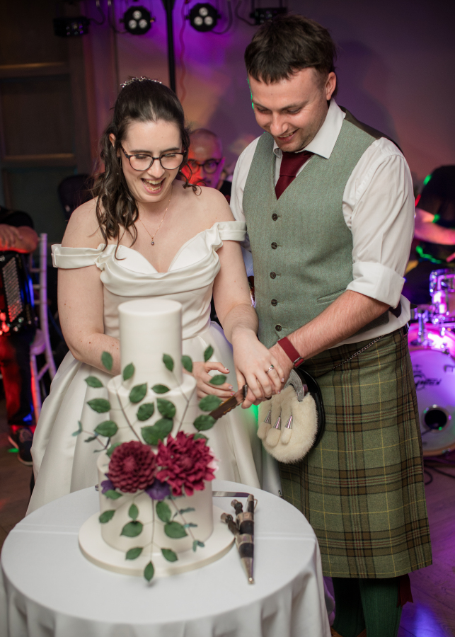 The bride and groom smile as they cut their wedding cake together during the evening reception. The bride wears an elegant off-the-shoulder white gown, while the groom wears a sage green waistcoat with a traditional Scottish tartan kilt. Their three-tier white cake is decorated with green vines and deep burgundy flowers, with a live band performing in the background.