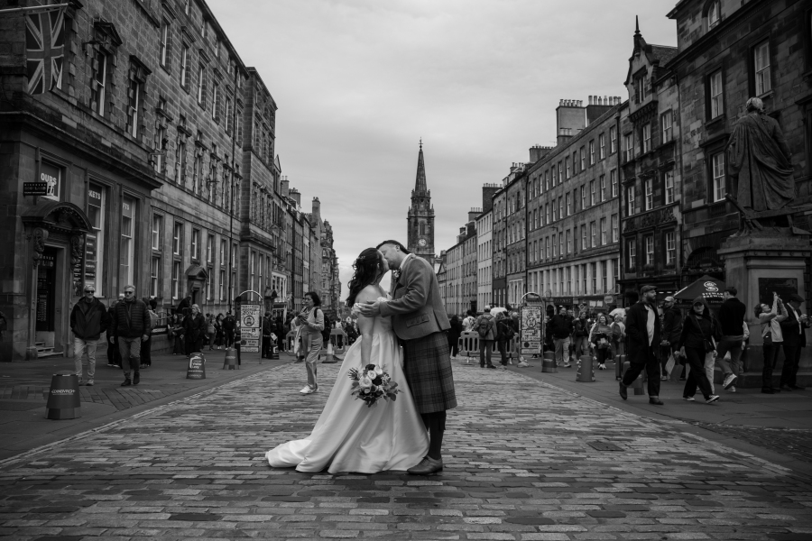 The bride and groom share a kiss in the middle of Edinburgh's historic Royal Mile, surrounded by bustling city life. The black-and-white photograph captures the bride in an elegant off-the-shoulder white gown holding a bouquet of white and burgundy flowers, while the groom wears a traditional Scottish tartan kilt with a sage green tweed jacket. The iconic cobbled street and historic architecture create a striking backdrop for their romantic wedding portrait.