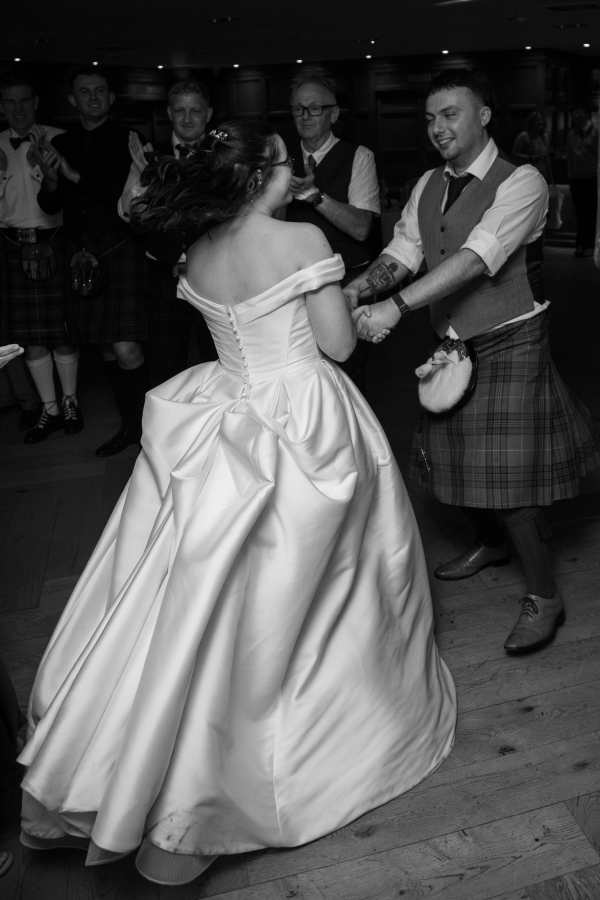The newlyweds share a joyful dance during their wedding reception, holding hands as guests clap around them. The bride wears an elegant off-the-shoulder satin ballgown with a dramatic bow detail at the back, while the groom wears a traditional Scottish tartan kilt. The black-and-white photograph captures the movement, happiness, and celebration of their first dance.