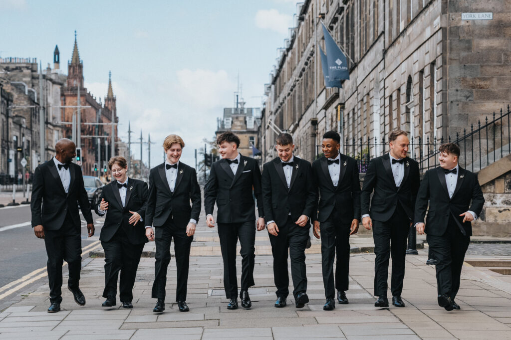 The groom and his groomsmen walking together through Edinburgh city centre in matching black tuxedos, sharing smiles and laughter before the wedding ceremony.