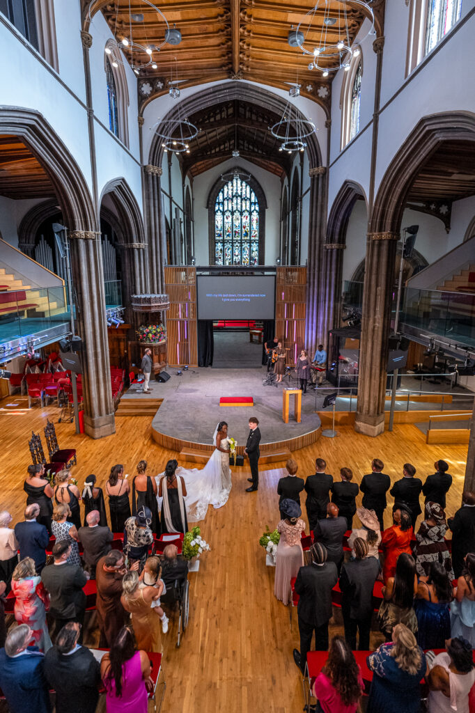 Bride and groom exchanging vows during a modern church wedding ceremony, surrounded by family and friends beneath the soaring arches and stained glass of Ps & Gs Church in Edinburgh.