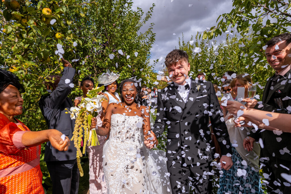 Bride and groom walking through a joyful confetti shower as family and friends celebrate after their wedding ceremony, with smiles, flowers, and sunshine all around.
