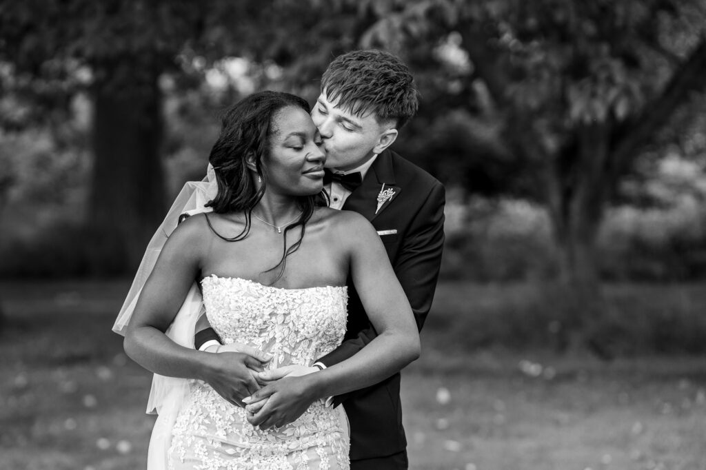 Black-and-white wedding portrait of the groom kissing the bride on the cheek as they embrace in a peaceful garden, capturing a tender moment during their wedding day.