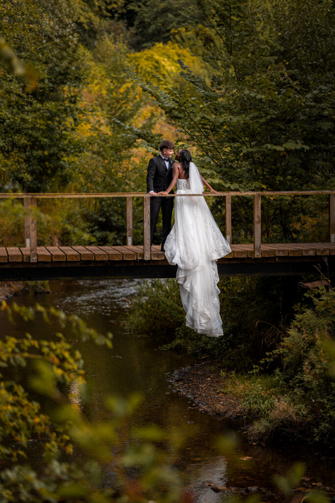 Bride and groom sharing a romantic moment on a wooden bridge over a peaceful stream, surrounded by lush woodland, during their outdoor wedding portrait session.