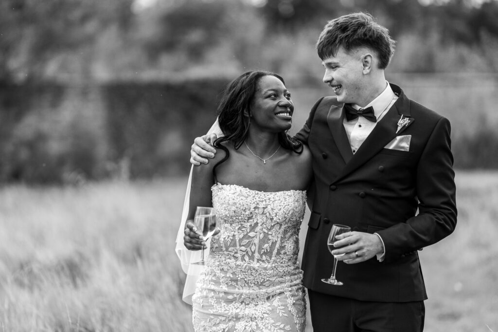 Black-and-white wedding portrait of the bride and groom smiling at each other while holding champagne glasses, sharing a joyful moment during their outdoor wedding celebration.