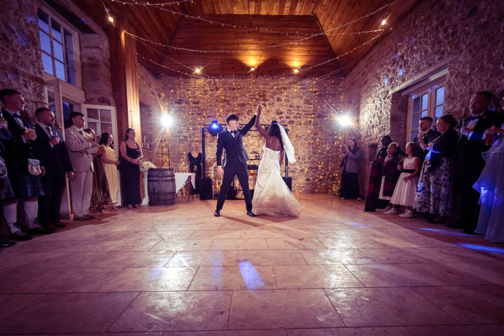 Bride and groom performing their first dance in a beautifully lit stone reception venue, surrounded by family and friends as they celebrate their wedding day.