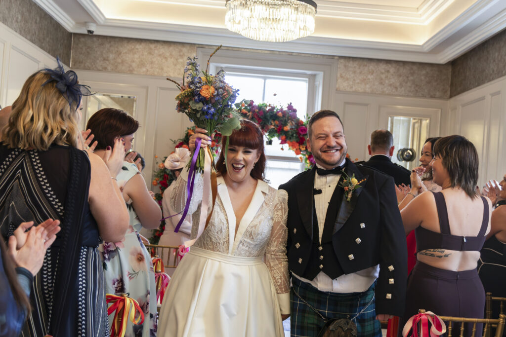 A joyful bride in a white lace gown and a groom in a traditional Scottish kilt walk down the aisle, smiling as guests applaud their wedding ceremony.