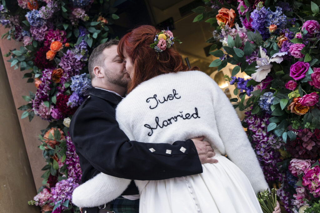 A groom in a black kilt jacket kisses his red-haired bride, who wears a white 'Just Married' sweater, as they embrace under a vibrant floral arch.