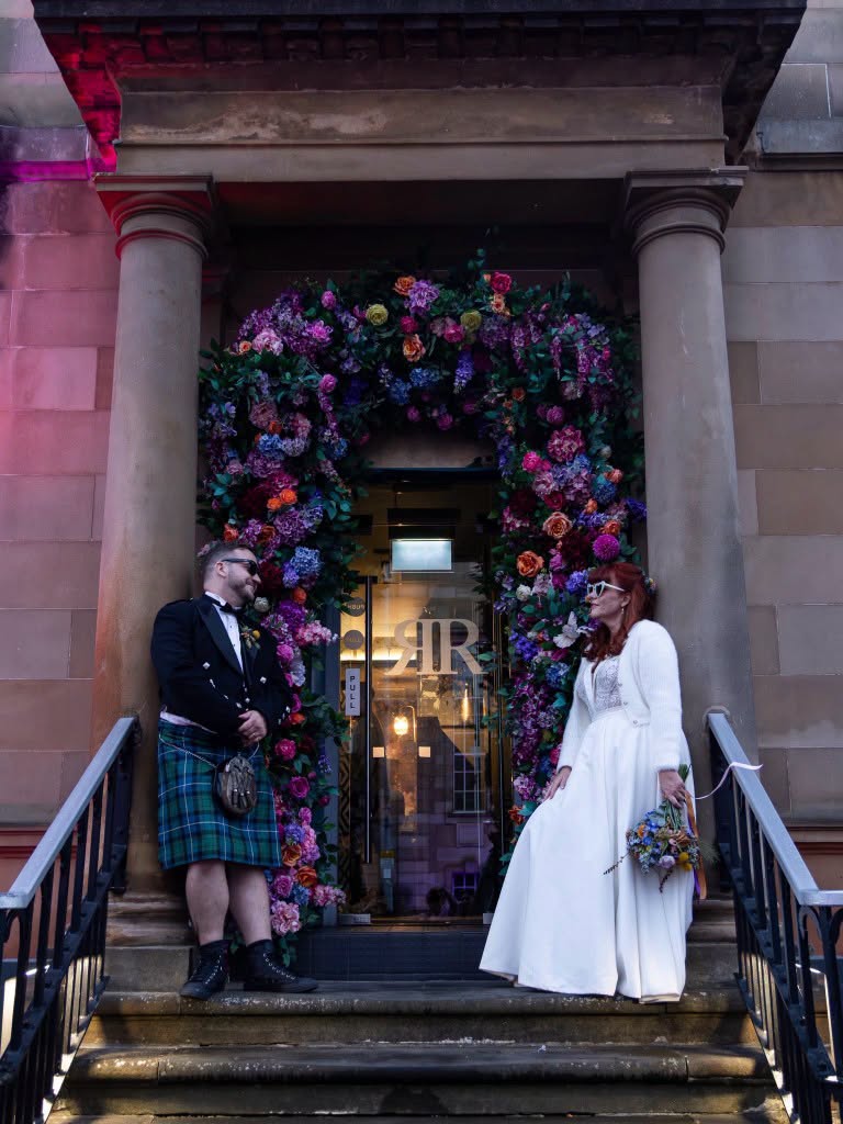 Bride and groom standing beneath a vibrant floral arch outside their wedding venue, sharing a joyful moment.